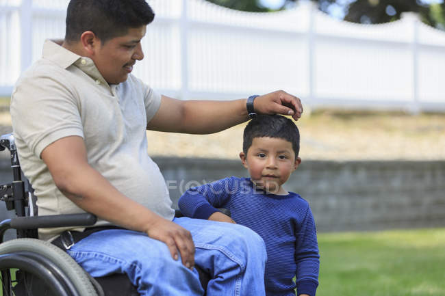 Homme hispanique avec lésion médullaire en fauteuil roulant avec son fils dans la pelouse — Photo de stock