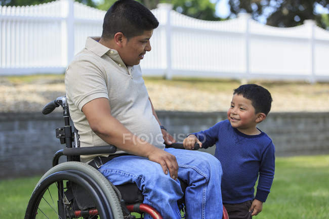 Homem hispânico com lesão medular em cadeira de rodas com seu filho no gramado — Fotografia de Stock