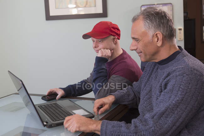 Padre con lesión de médula espinal e hijo con síndrome de Down con computadora portátil en casa - foto de stock