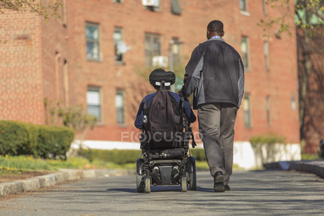 Hombre afroamericano con parálisis cerebral usando su silla de ruedas eléctrica afuera con su asistente de cuidado personal - foto de stock