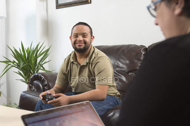 Heureux homme afro-américain avec le syndrome de Down en utilisant le contrôleur de jeu et la mère en utilisant un ordinateur portable à la maison — Photo de stock