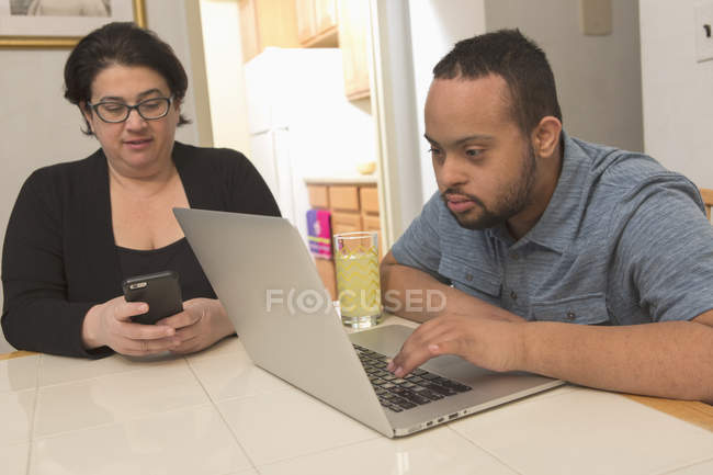 Heureux homme afro-américain avec le syndrome de Down en utilisant un ordinateur portable avec la mère à la maison — Photo de stock