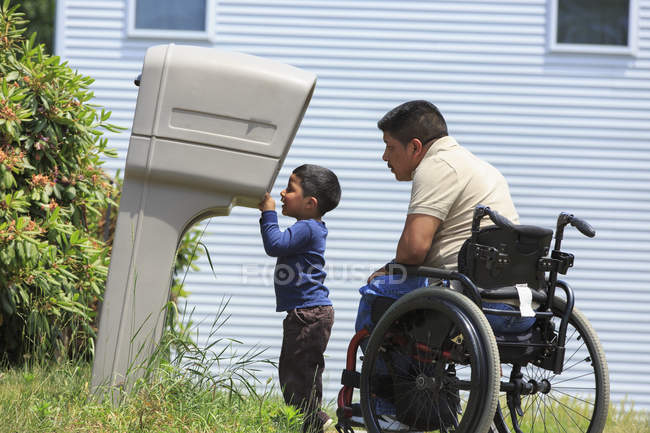 Hombre hispano con lesión de médula espinal en silla de ruedas con su hijo mirando en un buzón - foto de stock
