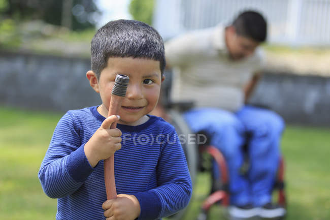 Портрет латиноамериканца, держащего в руках шланг, и его отца в инвалидной коляске с травмой спинного мозга на заднем плане — стоковое фото