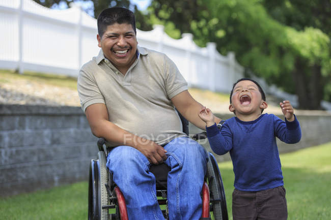 Латиноамериканец с травмой спинного мозга в инвалидном кресле со смехом сына на газоне — стоковое фото