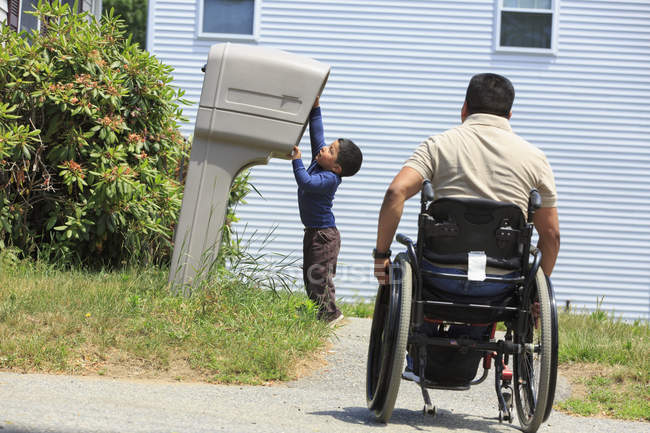 Hombre hispano con lesión de médula espinal en silla de ruedas con su hijo mirando en un buzón - foto de stock