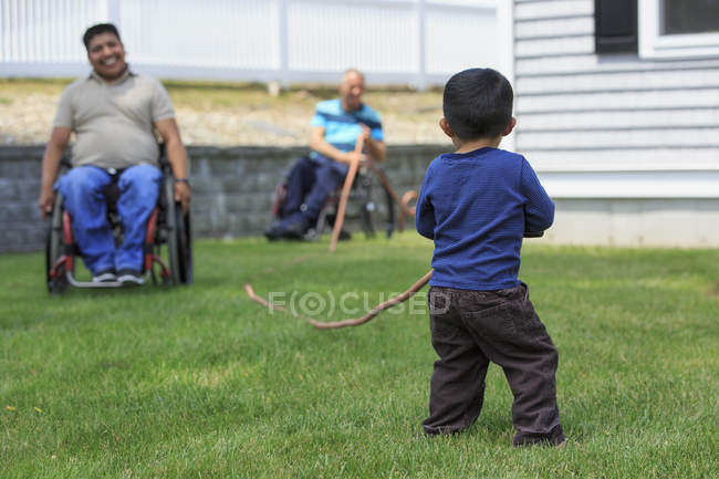 Garçon jouer avec tuyau accompagné par des hommes avec blessures de la moelle épinière sur les fauteuils roulants dans la pelouse — Photo de stock