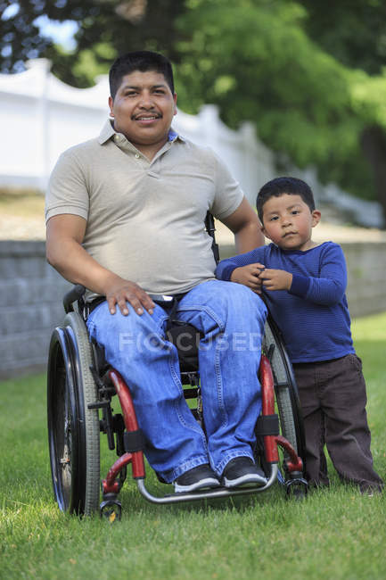 Retrato del hombre hispano con lesión de médula espinal en silla de ruedas con su hijo en el césped - foto de stock