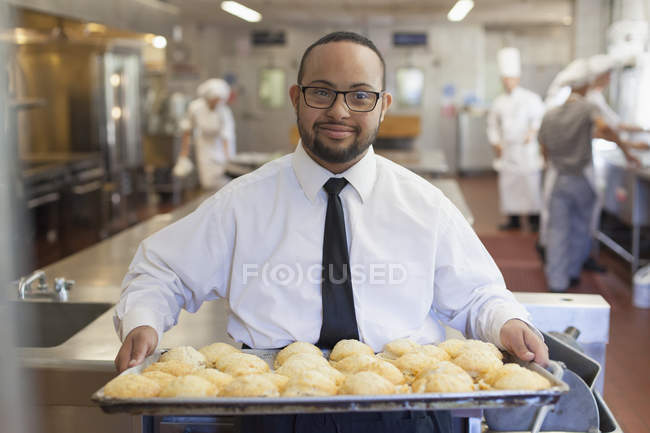 Афроамериканец с синдромом Дауна в качестве шеф-повара держит поднос печенья на коммерческой кухне — стоковое фото