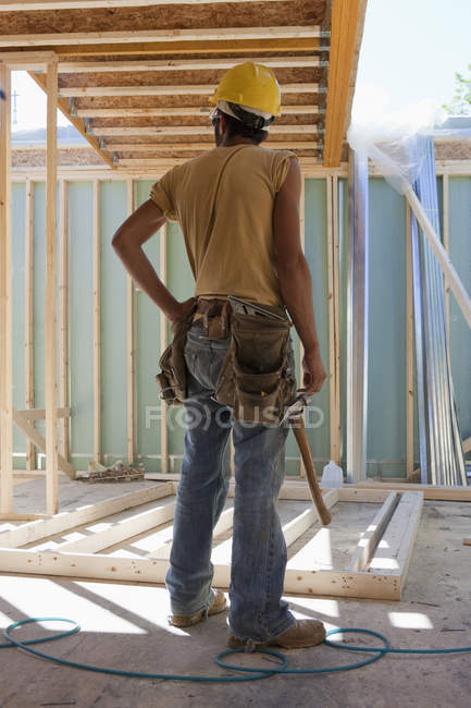 Falegname ispanico con un martello in piedi accanto al telaio di una casa in costruzione — Foto stock