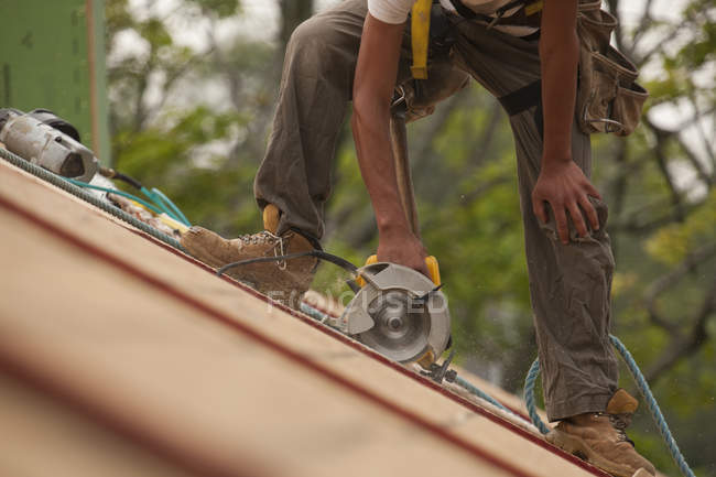 Charpentier hispanique utilisant une scie circulaire sur le panneau de toit d'une maison en construction — Photo de stock