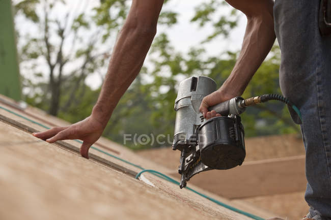 Charpentier hispanique utilisant un pistolet à clous sur le panneau de toit d'une maison en construction — Photo de stock