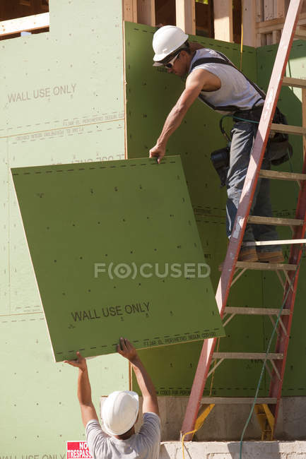 Hispanic carpenters pulling exterior sheathing up ladder at a house under construction — Stock Photo