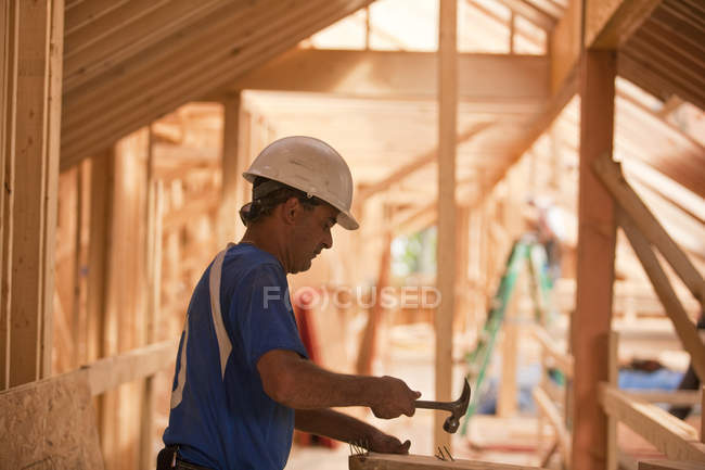 Carpinteiro hispânico martelando pregos a bordo em uma casa em construção — Fotografia de Stock