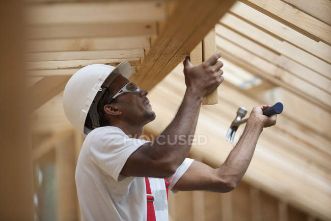 Carpintero hispano martillando tablas sobre techos en una casa en construcción - foto de stock