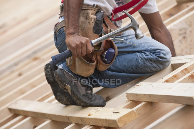 Carpintero con un martillo y correa de herramientas en el techo de una casa en construcción - foto de stock