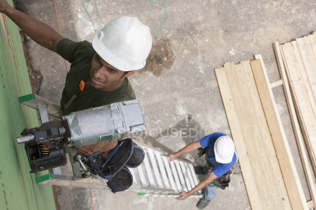 Hispanic carpenter using nail gun on top of ladder being steadied by another carpenter — Stock Photo