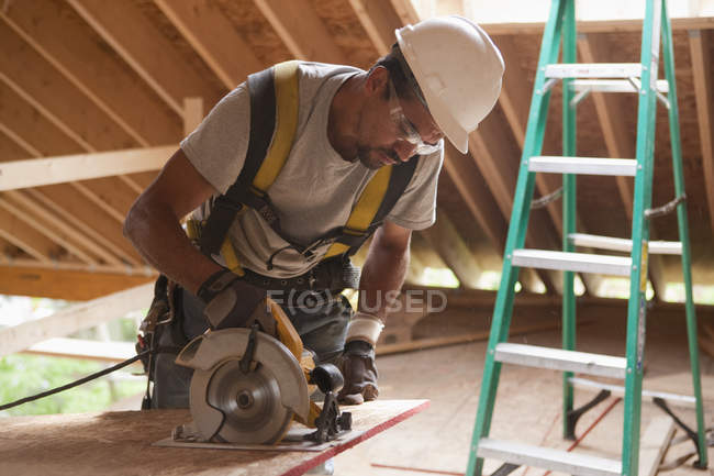 Carpinteiro hispânico usando uma serra circular no painel do telhado em uma casa em construção — Fotografia de Stock