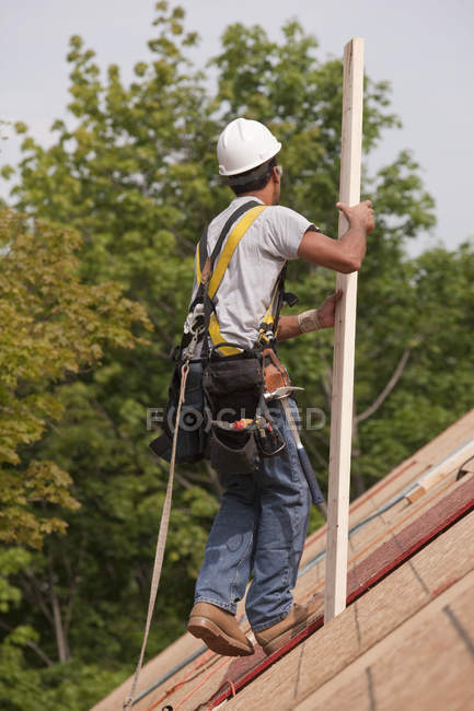 Carpintero hispano llevando tablón en el techo de una casa en construcción - foto de stock
