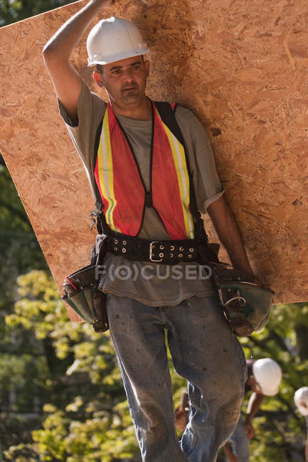 Carpintero que lleva un tablero de partículas en un sitio de construcción - foto de stock