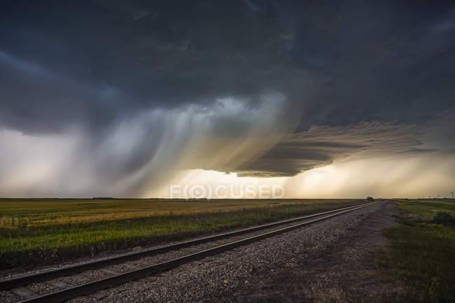Nuages de tempête foncés au-dessus des voies ferrées dans un champ où la pluie tombe au loin ; Marquis, Saskatchewan, Canada — Photo de stock