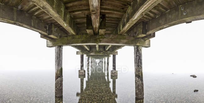 Нижняя часть пирса Crescent Beach отражена в спокойной воде в туманный день; Суррей, Британская Колумбия, Канада — стоковое фото