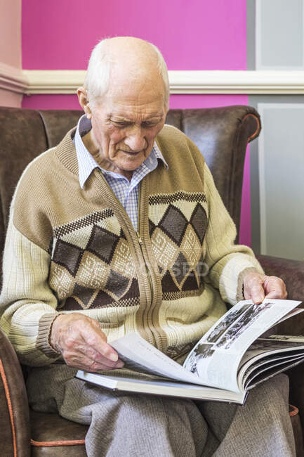 Hombre mayor sentado en una silla mirando un libro sobre la Segunda Guerra Mundial; Hartlepool, Condado de Durham, Inglaterra - foto de stock