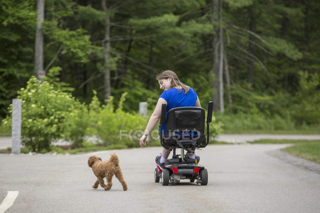 Mujer joven con parálisis cerebral jugando con su perro mientras está sentada en su scooter - foto de stock