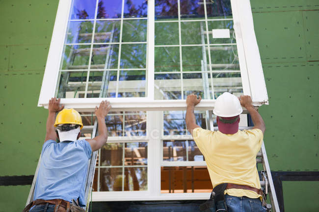 Menuisiers positionnant un grand cadre de fenêtre sur une maison en construction — Photo de stock
