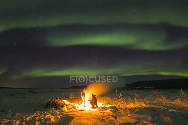 Mantenerse caliente junto a una fogata en el río Delta mientras observa la aurora boreal en una noche fría; Alaska, Estados Unidos de América - foto de stock