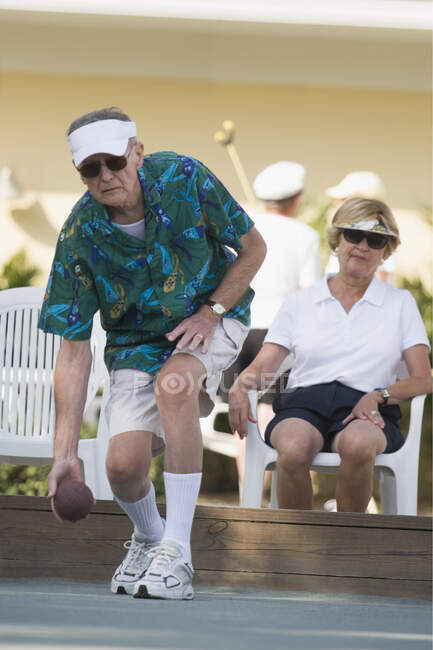 Старший мужчина играет в бочче-бол со старшей женщиной, сидящей на стуле позади него — стоковое фото