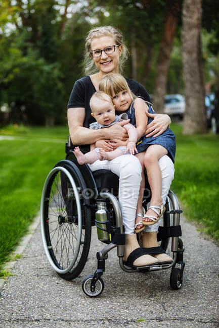 Eine querschnittsgelähmte Mutter trägt ihre Töchter auf dem Schoß, während sie an einem warmen Sommernachmittag im Freien im Rollstuhl für die Kamera posiert: Edmonton, Alberta, Kanada — Stockfoto
