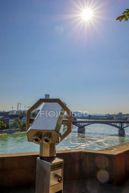 Binocolo lungo il lungomare che si affaccia sul fiume Reno con ponte e un raggio di sole nel cielo blu; Basilea, Basilea Città, Svizzera — Foto stock