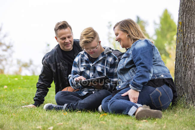Ein junger Mann mit Down-Syndrom lernt mit seinem Vater und seiner Mutter ein neues Programm auf einem Tablet, während er an einem warmen Herbstabend in einem Stadtpark Gesellschaft genießt: Edmonton, Alberta, Kanada — Stockfoto