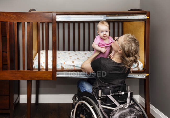 A paraplegic mother lifting a baby from a customized side-opening crib that allows her to put her baby down for a nap from her position in a wheelchair: Edmonton, Alberta, Canada — Stock Photo