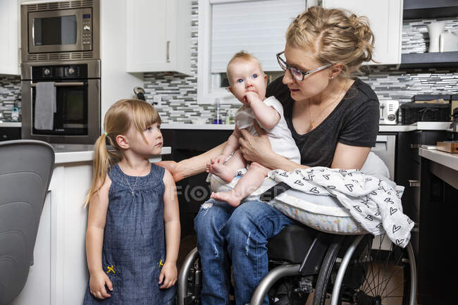 Параплегічна мама в інвалідному візку розмовляє з дочкою і тримає своє немовля на колінах під час роботи на кухні; Едмонтон (Альберта, Канада). — стокове фото