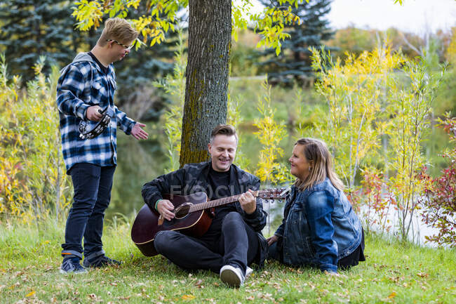 Ein junger Mann mit Down-Syndrom spielt Tamburin, während sein Vater Gitarre spielt und seine Mutter mitsingt, während sie an einem warmen Herbstabend in einem Stadtpark Gesellschaft genießt: Edmonton, Alberta, Kanada — Stockfoto