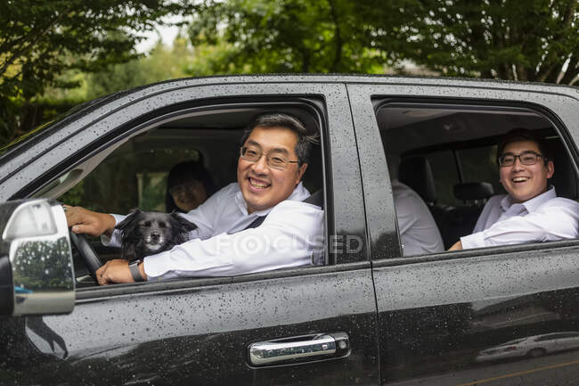 Promenade en famille dans la voiture avec le chien, le père conduisant et les enfants adultes sur le siège arrière, regardant la caméra et souriant ; Langley, Colombie-Britannique, Canada — Photo de stock