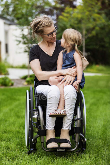Una madre parapléjica sosteniendo a su niña en su regazo mientras está sentada en su silla de ruedas en su patio delantero en una cálida tarde de verano: Edmonton, Alberta, Canadá. - foto de stock