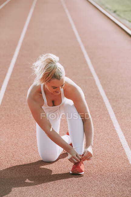 Mujer arrodillada para atar su zapato de correr para prepararse para correr en una pista; Wellington, Nueva Zelanda - foto de stock