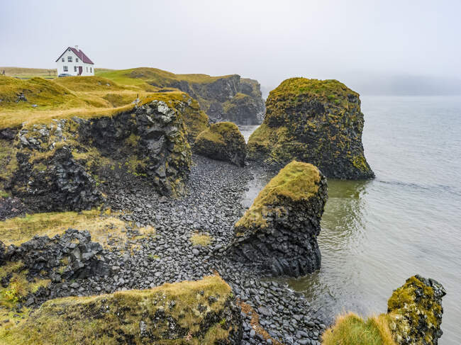 Robusta costa de la península de Snaefellsnes con una casa solitaria a lo largo de los acantilados en la niebla; Snaefellsbaer, Región occidental, Islandia - foto de stock