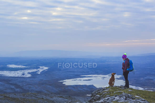 Lone woman hiker with knit hat and dog looking at each other on a cliff edge overlooking lakes in the distance on a cloudy evening in winter, Burren National Park; County Clare, Ireland — Stock Photo