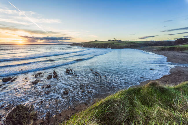 Puesta de sol de verano sobre la cala de la playa de Inch con olas que se estrellan en el mar y hierba en primer plano; Condado de Cork, Irlanda - foto de stock