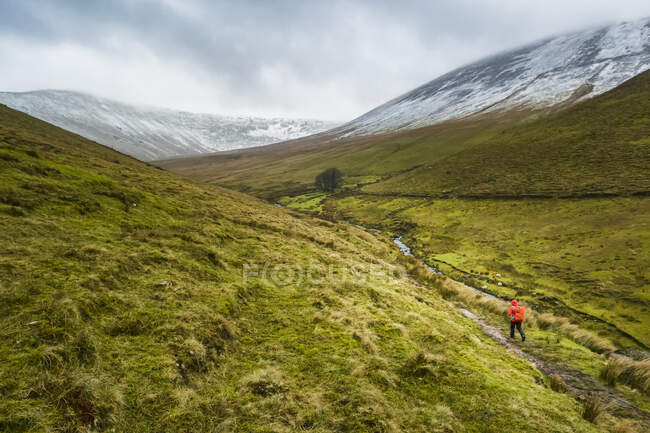 Einsame Wanderin in rotem Regenmantel mit rotem Rucksack auf einem Talweg, der an einem bewölkten Wintertag in die schneebedeckten Berge führt, Galty Mountains; County Tipperary, Irland — Stockfoto