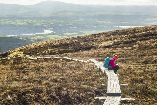 Senderista solitaria con una mochila sentada en un sendero de madera que lee un mapa en una montaña en un día soleado con un río y campos al fondo; Killaloe, Clounty Clare, Irlanda - foto de stock