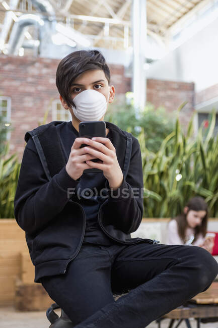 Garçon à la maison avec un téléphone intelligent portant une masse protectrice pour se protéger contre la COVID-19 pendant la pandémie mondiale de coronavirus, et une fille en arrière-plan ; Toronto, Ontario, Canada — Photo de stock