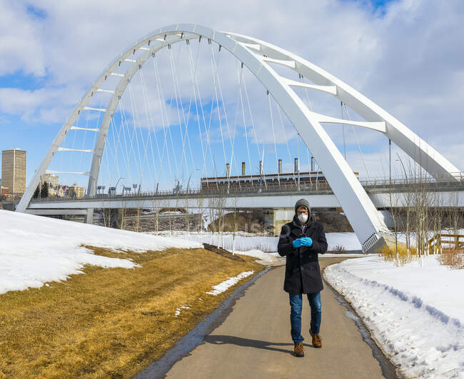 Man wearing a mask and gloves walking on a path outside using his smart phone during the Covid-19 world pandemic; Edmonton, Alberta, Canada — Stock Photo