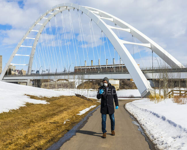 Uomo che indossa una maschera e guanti camminando su un percorso esterno utilizzando il suo smartphone durante la pandemia mondiale Covid-19; Edmonton, Alberta, Canada — Foto stock