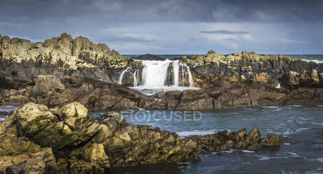 Robusta costa y agua que fluye sobre rocas; Bamburgh, Northumberland, Inglaterra - foto de stock