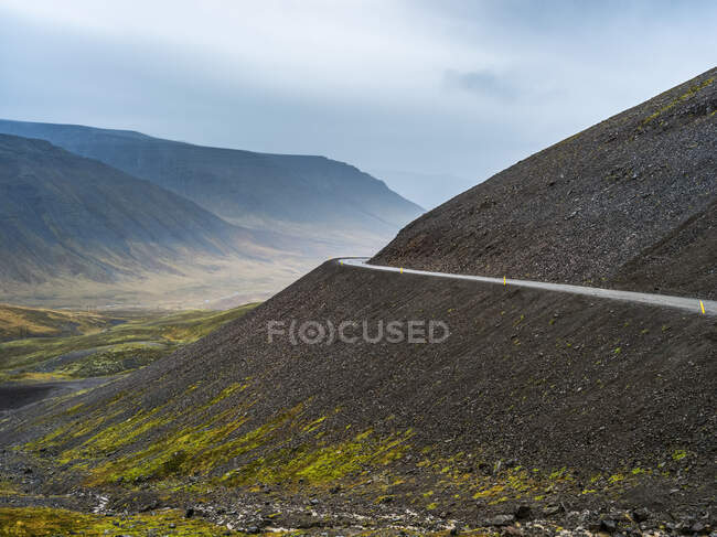 Camino que va alrededor de una curva en una ladera con una vista de un valle y montañas bajo un cielo nublado; Westfjords, Islandia - foto de stock
